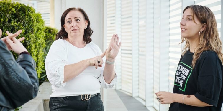 A photo of Centre for Inclusive Design’s team. One team member is teaching the two others in the photo Auslan (Australian sign language).